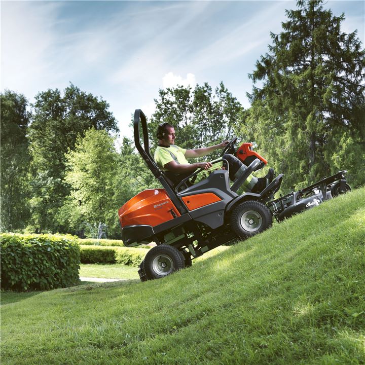 Oranje Husqvarna zitmaaier bestuurd door een man met gehoorbescherming op een groene grashelling onder een blauwe lucht.